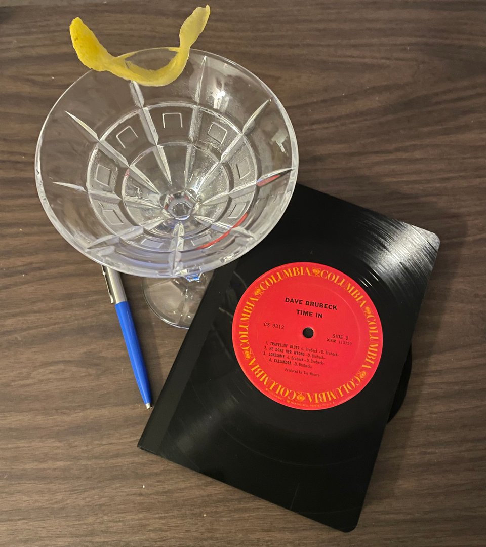 Objects on a coffee table. A martini with a lemon twist. A blue pen. And a notebook with an old vinyl record as the cover