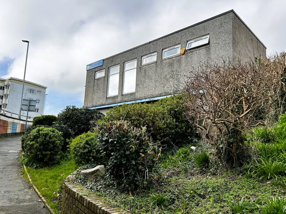 A rough concrete wall of a squarish building. It has two full length windows and four narrow high windows. In the foreground, a slope up from an underpass has some sad, ivy-throttled shrubs on it.