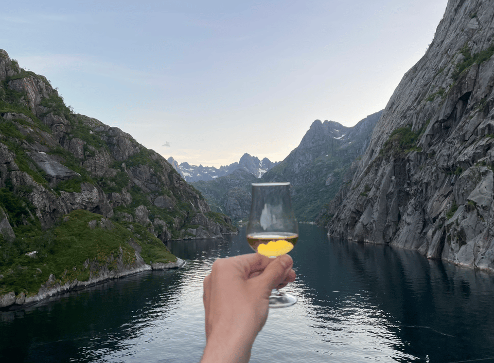 A hand holding a small glass of amber fluid toward a range of mountains jutting from the water