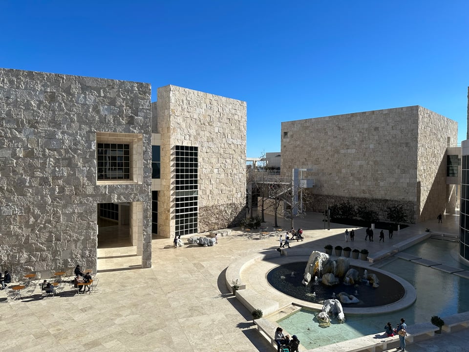 Photo of several buildings of the Getty Center in LA. They are stone square facades in bright noon sun, with a blue sky in the background. A fountain is in the lower right corner, with a duck.