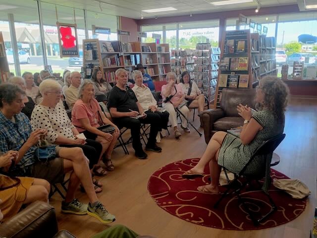 A seated woman in a dress shown from the side and behind, sitting before an audience of people in folding chairs with shelves of books around and behind them.