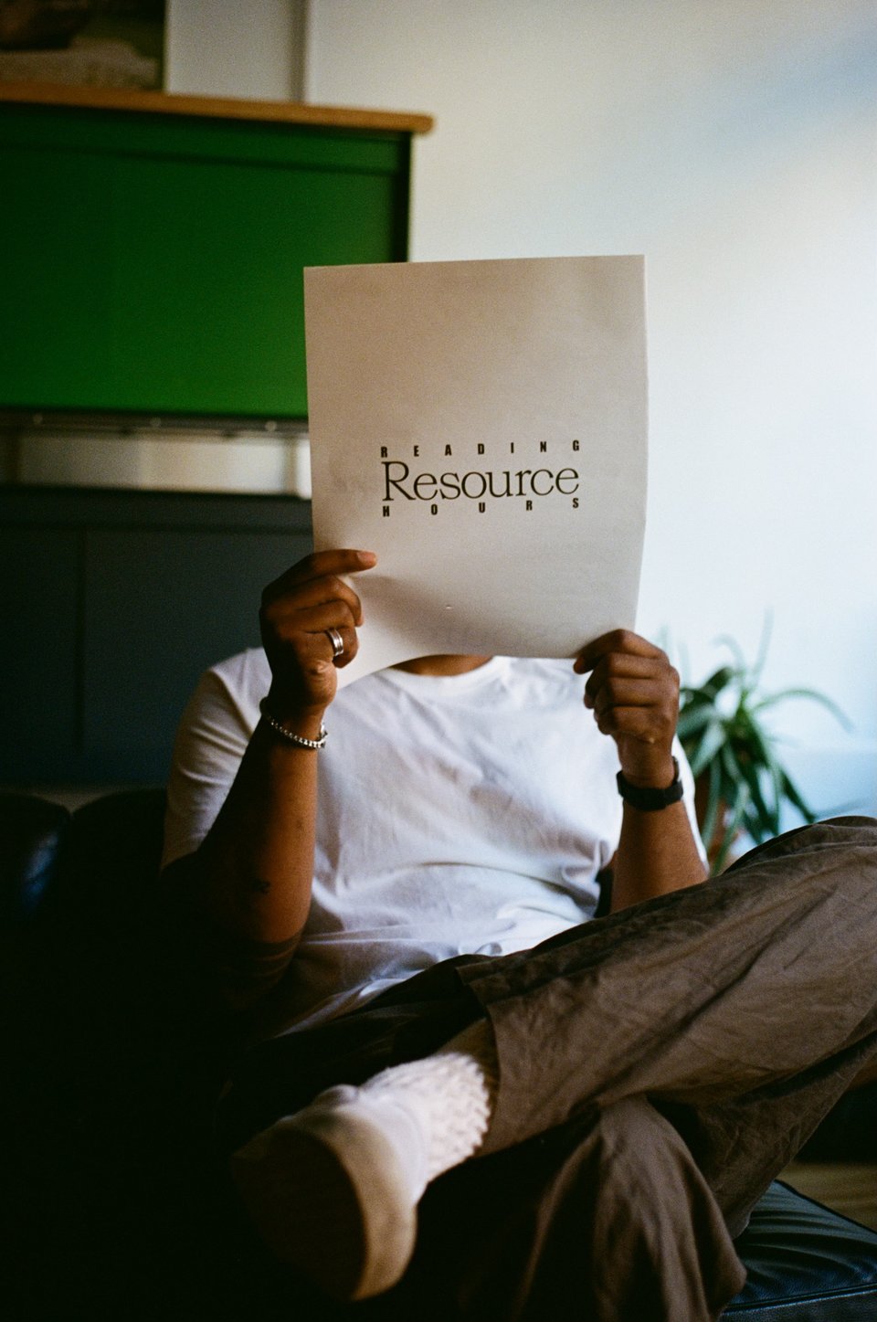 [A person sits in a chair and reads a broadsheet with Resource Reading Hours on the front of it.]