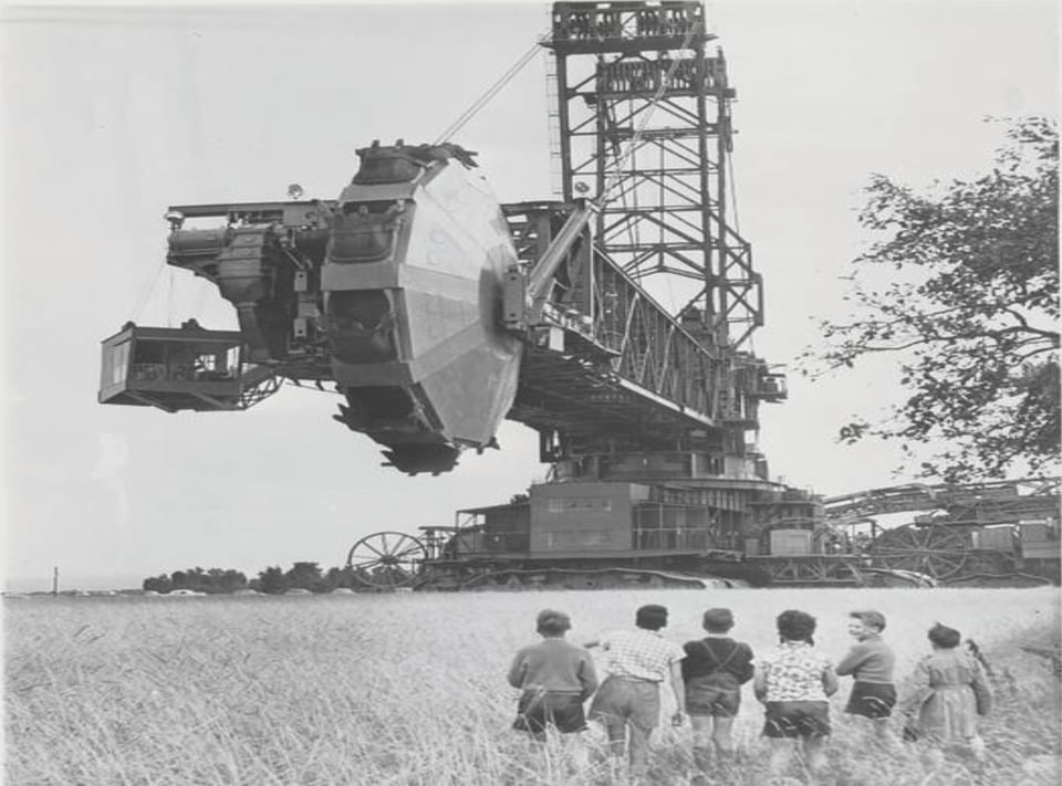 In a field of wheat, six children look up at a gigantic machine that looks like a metal bridge with a massive mill wheel at the end of it.