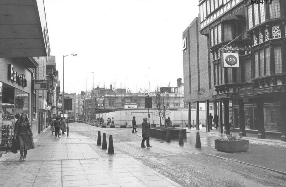 Photo of Exeter High Street. There is a building site in the middle of the shot. To its right is a brick-clad brutalist building that lines up nicely with the sixteenth century wooden merchant's house next to it. Both the brutalist and Tudor buildings have C&A signs hanging on them.