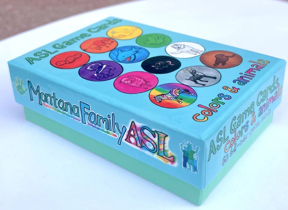 Commercial photo of a card game box. The box text is done in a multicolor, handmade script, and reads "Montana Family ASL, ASL game cards, colors & animals." The box is blue, with multicolored circles on the front with drawings of various animals inside.
