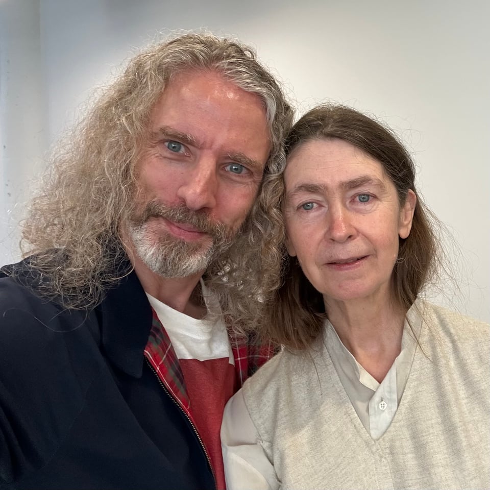 Photo of a man with long, curly hair and a beard, in a blue jacket, and a woman with straight brown hair, in a light beige dress and an off-white buttondown, smiling for the camera. Their hair is touching and they are concealing thoughts about art.