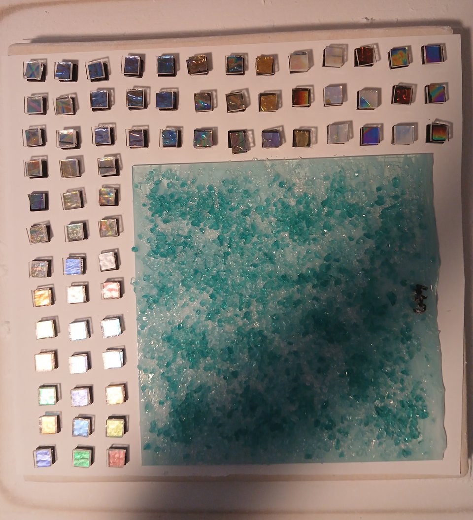 a kiln shelf with a sheet of blue glass covered in blue frit and surrounded by 69 square stacks of dichroic glass sandwiched between black and clear