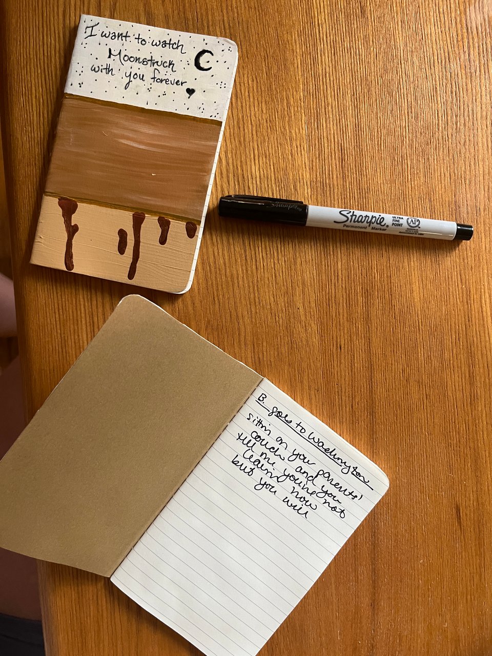 small homemade notebooks on a wooden table. the cover of one says "I want to watch Moonstruck with you forever" and features gold paint and dark gold dripping wax. the other is open and shows the song title "B. goes to Washington" and the lyric "sittin on your parents' couhc and you tell me you're not leavin now but you will" in black Sharpie cursive.