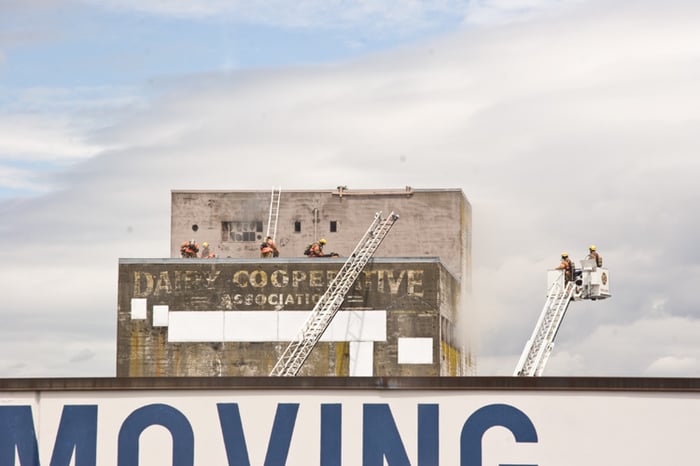 A photo of a concrete-brick building with faded dairy signage, firefighters on ladders about the roof, another cherry-picker type ladder extended to the side. The front of the building and front of the image is blocked off by a large sign that says "MOVING" painted on a closer building, the sky is blue washed grey.