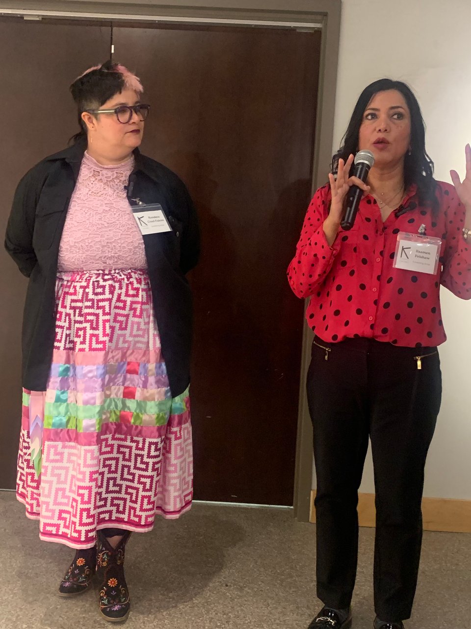 Two artists speaking about their work, on left the author in a pink ribbon skirt and on the right Ilaamen Pelshaw in a polka dot top holding a microphone