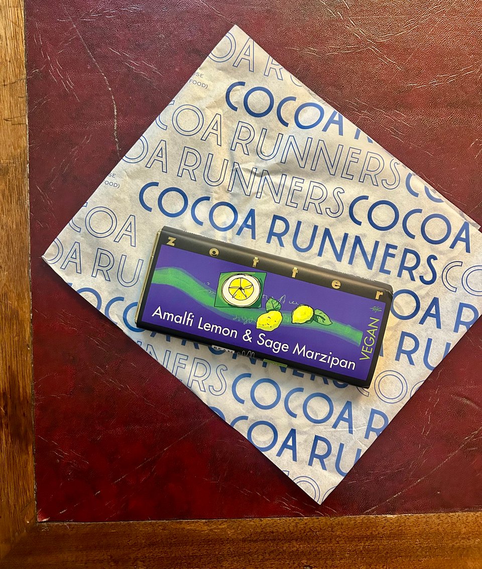 A bar of Zotter Amalfi Lemon and Sage Marzipan is on some waxed paper with Cocoa Runners branding on it. Everything is on a dark red leather topped desk. Image by Rowan Ambrose.