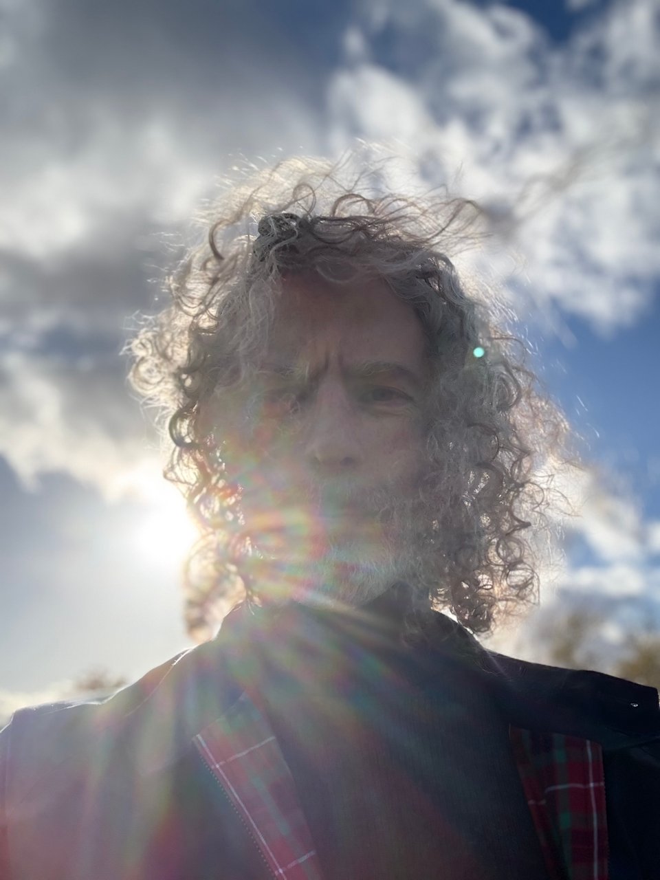 selfie of white man with long curly hair, with lens flare from sun behind him obscuring most of his features; he's wearing a dark turtleneck and a navy Baracuta Harington G9 jacket
