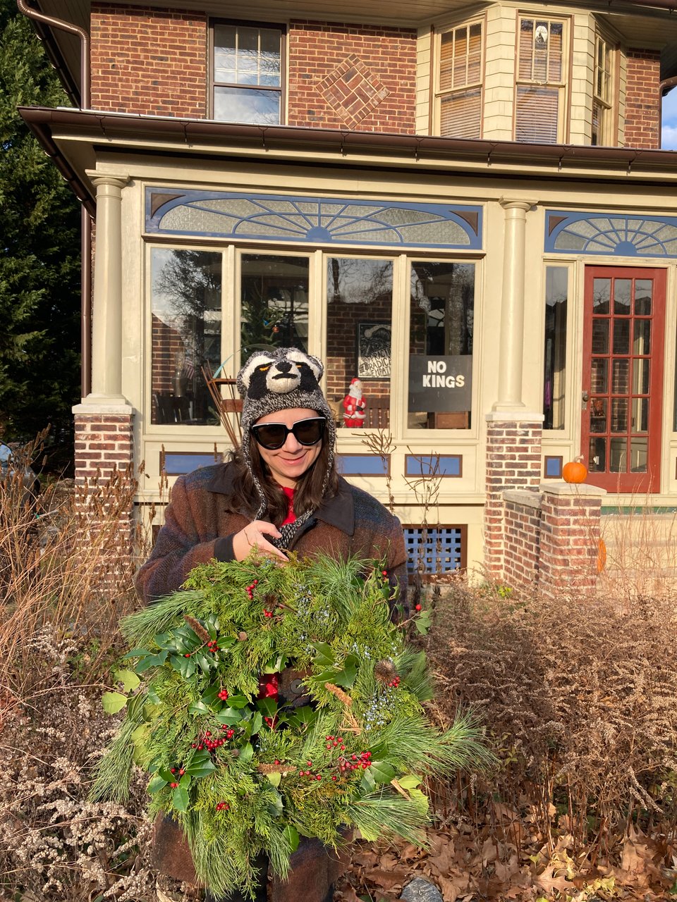 Woman in raccoon hat holding a native plant holiday wreath standing in native plant garden in front of a house.