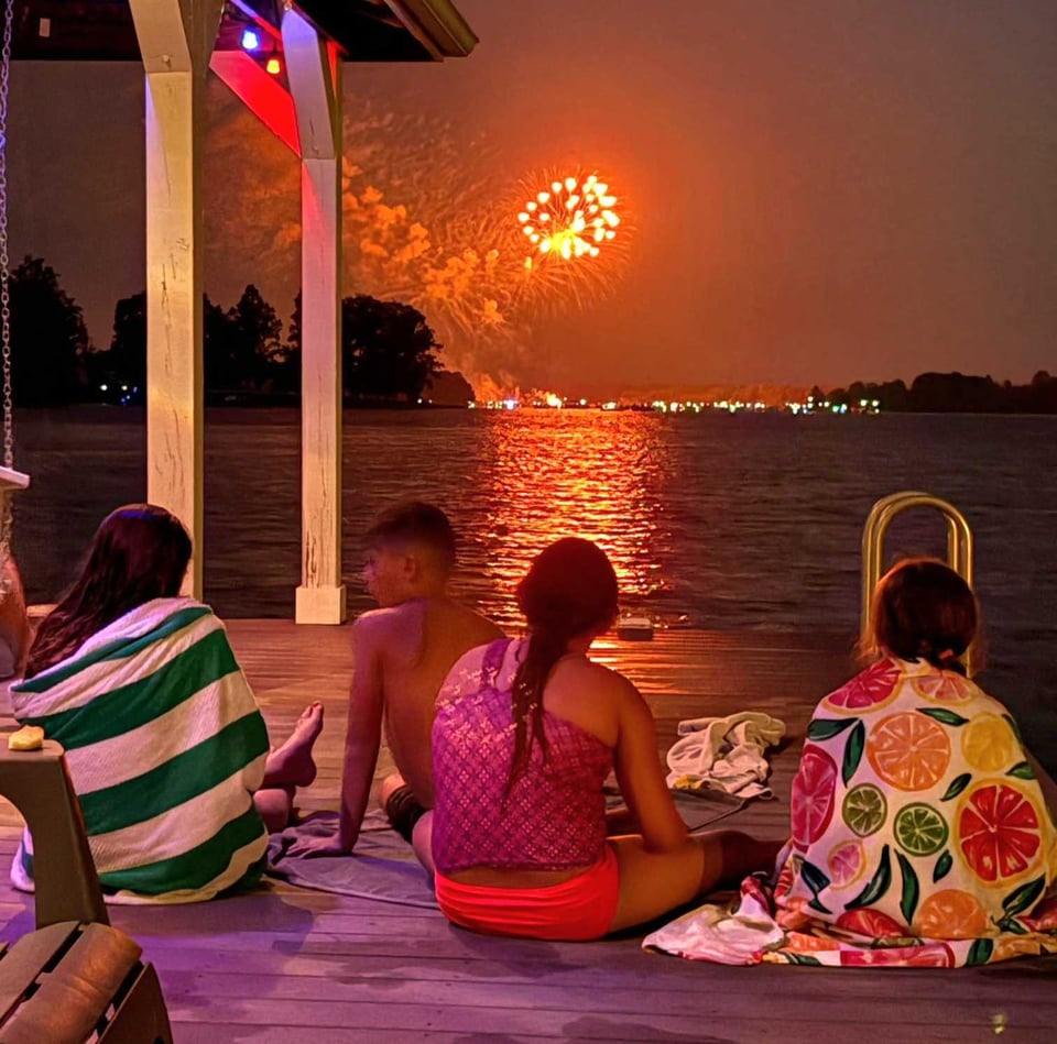 Fireworks over a lake view from a dock. Kids watch the fireworks wrapped in towels.