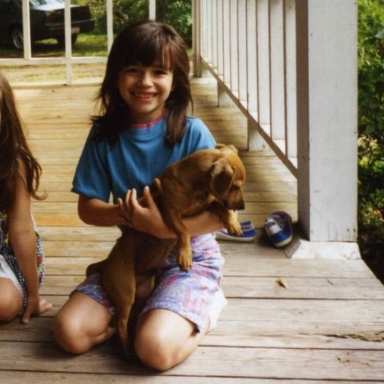 A smiling young brunette girl sits on a porch holding a small brown dog