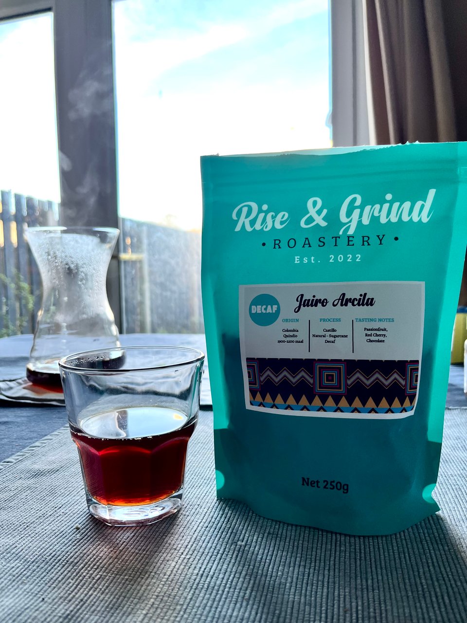 A turquoise bag of Jairo Arcila decaffeinated coffee from Rise and Grind is sitting on a table by a big window. There's a glass Hario coffee carafe and a glass, both with filter coffee in also on the table. Image by Rowan Ambrose.