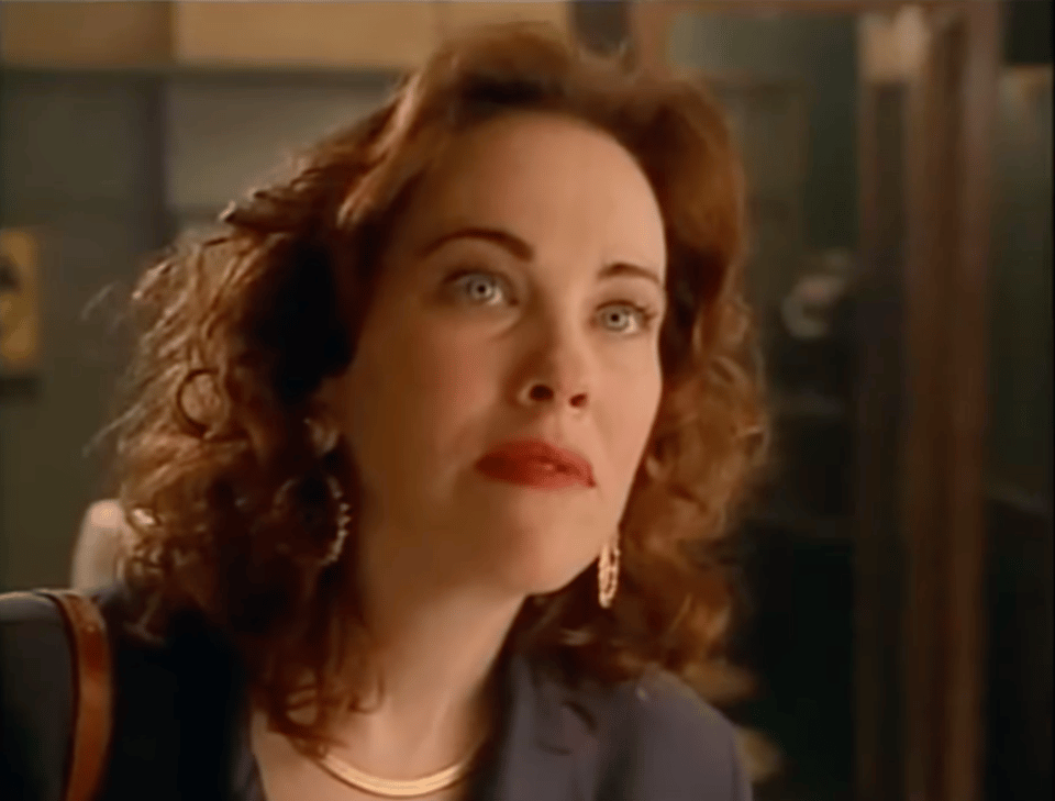 Catherine O'Hara, dressed in an olive power suit with a copper chain and earrings, looking up bemused at something on a wall (some photos of public hangings, for example)