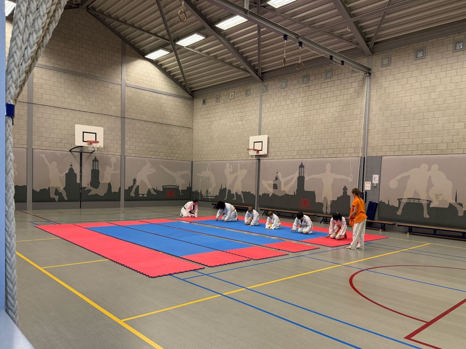 In a large grey gymnasium, six children in karate suits kneel down on the ground and help put all the mats together while the trainer looks on.
