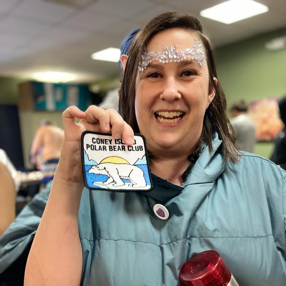 I'm wearing a blue-grey robe and smiling while holding up my polar bear club patch. I have face jewels on. They look... not good.