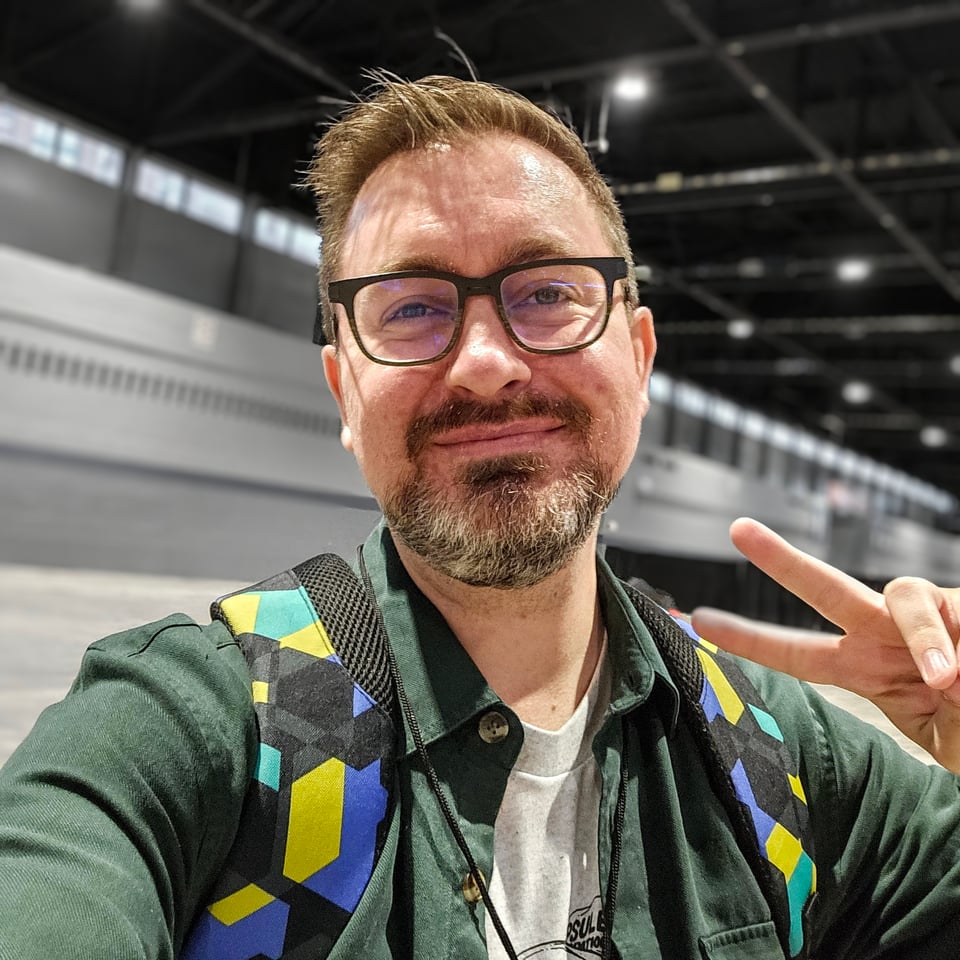 A man smiling in a big open convention center giving a peace sign.