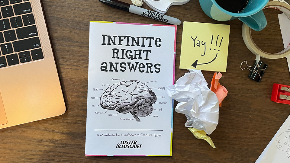 An image of a messy desk with a laptop, office supplies, and a zine entitled "Infinite Right Answers." The cover shows a brain on it.