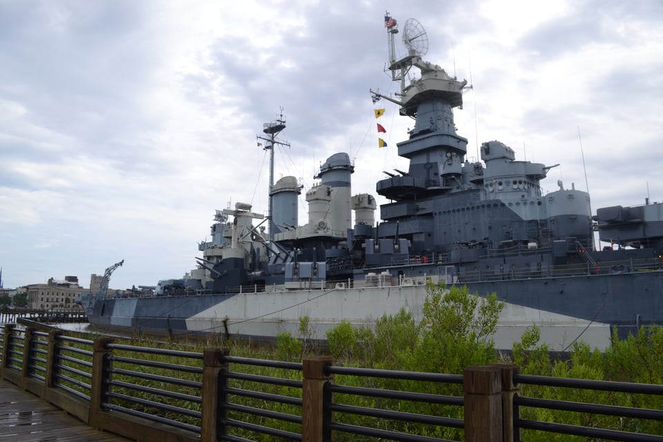 The side of a battleship painted in gray and creme, with the radar disk and superstructure visible. In the foreground is a fence with wood pillars and metal rails and there is water between the two.