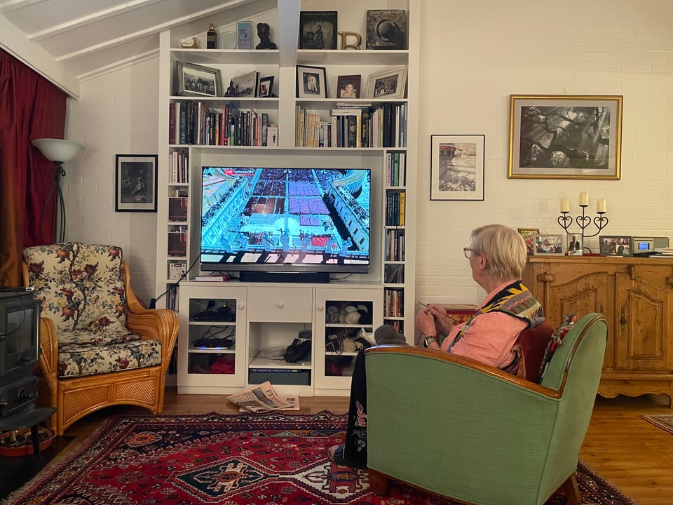 A TV in the middle of the living room screen shows the view of St Peters square in Rome for the Pope's funeral. A woman sits in a chair, watching the TV and knits.