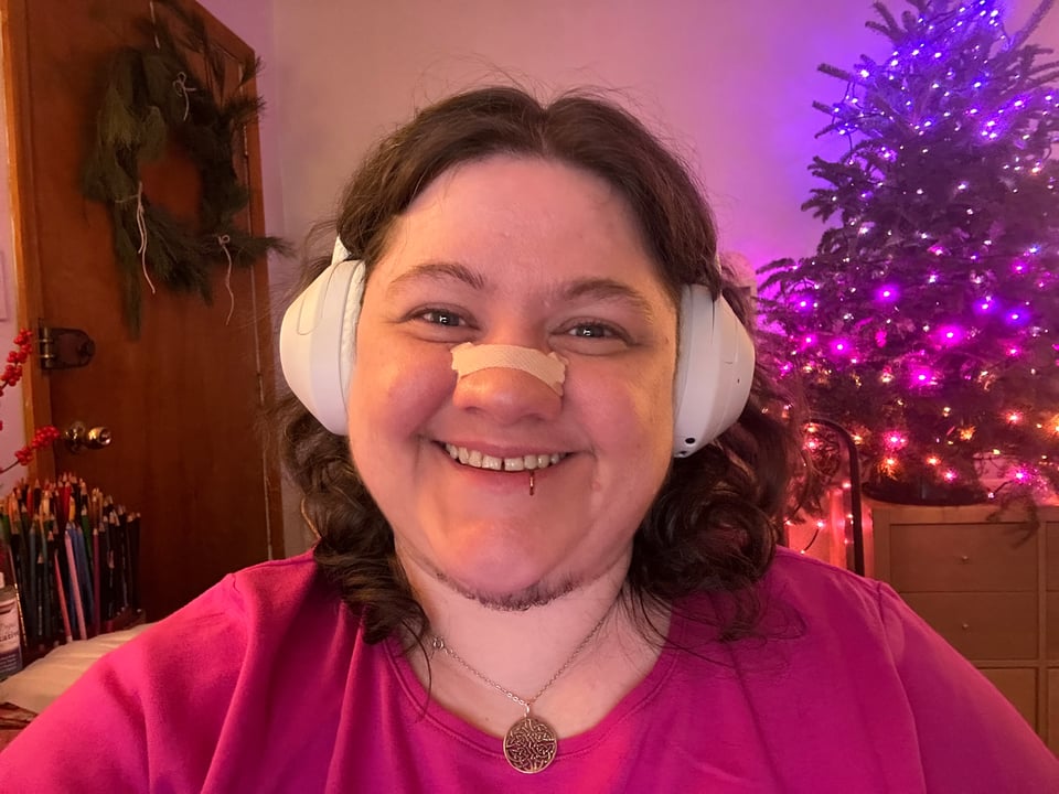 A photo of Jessi (a fat, white person with brown wavy hair and a gap-toothed grin) smiling at the camera. Behind them, a small pine tree is decorated with bright, twinkling lights in hues of pink and purple, and there's a wreath handing on their front door. The mood is very festive and cheery!