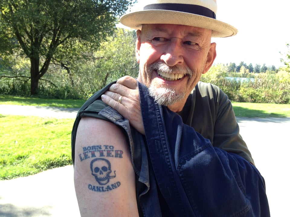 Man with gray beard pulls up his sleeve to reveal the tatto, "Born to letter", with a scull smoking a joint, and "Oakland" underneath.