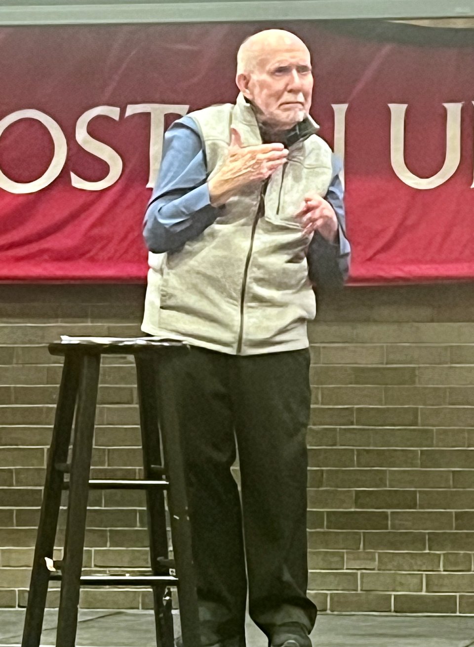 Patrick Graybill, a bald, elderly white man in casual clothes, stands on a stage. He is in the midst of signing a poem in ASL, frowning with emotion.