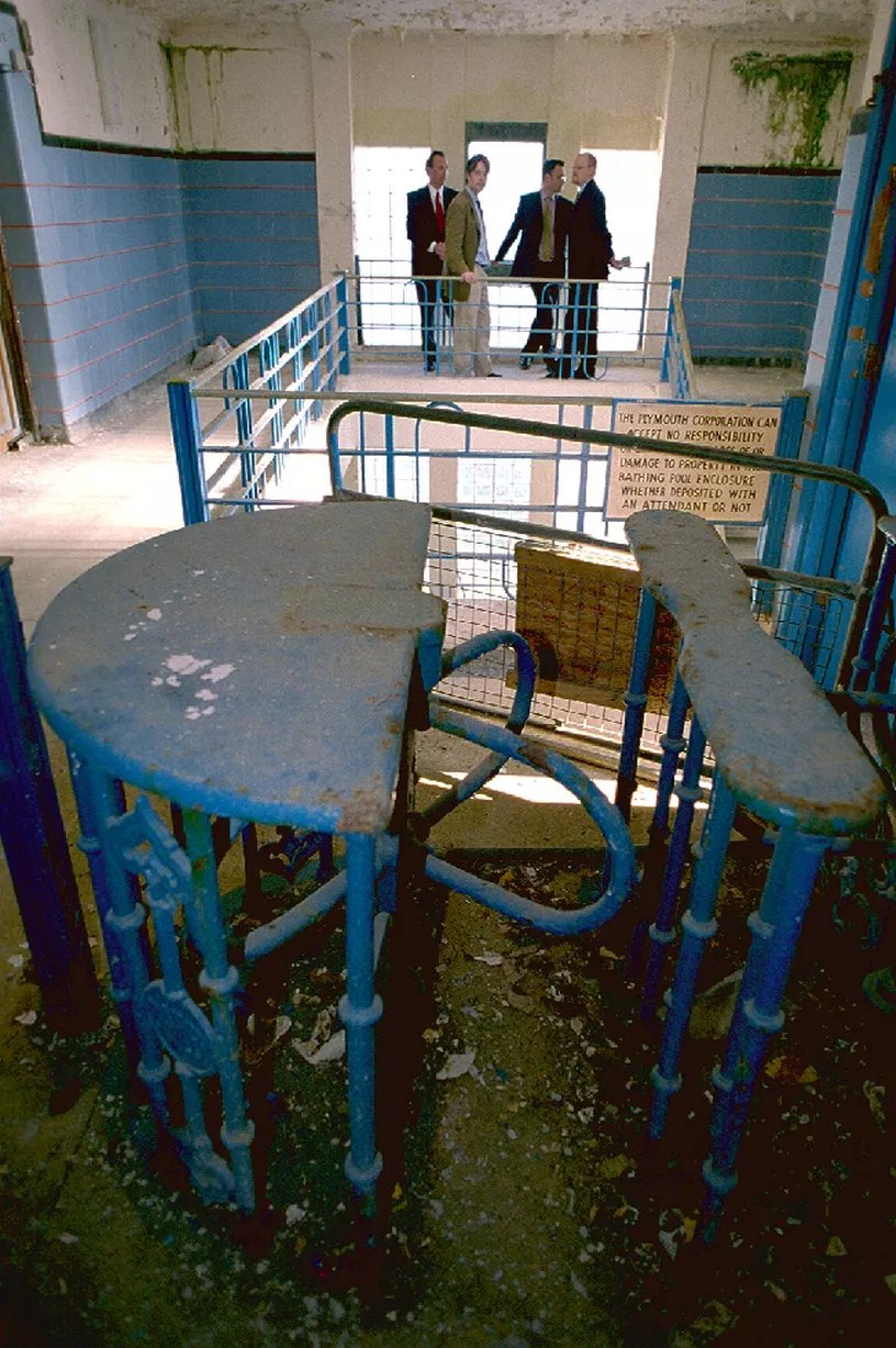 A tiled stairwell with a large iron turnstile in the foreground. A group of men are standing by the tall windows at the far side. The ceiling plaster is hanging in pieces, and there is green algae growing in the corners. Everything is covered with a layer of filth.