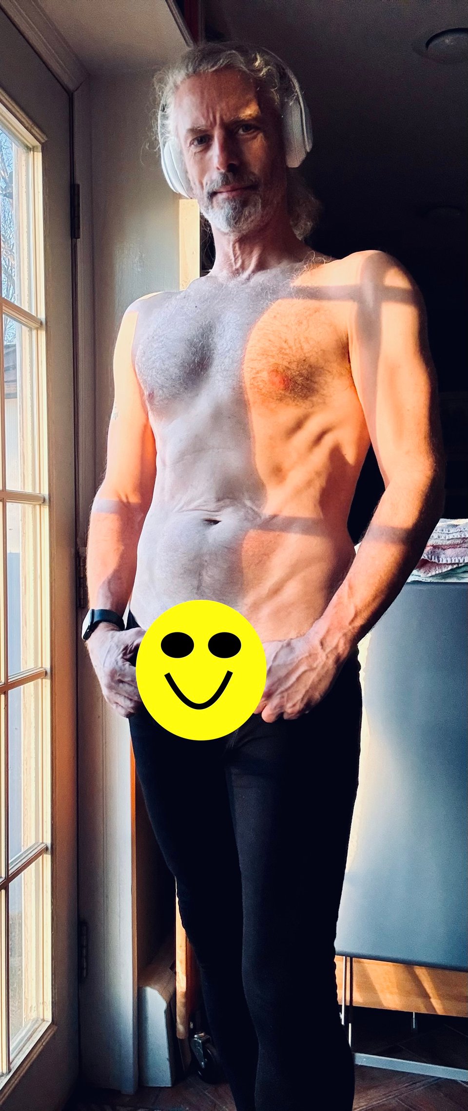 photo of shirtless white man in black tights, thumbs looped around waistband. There's a big smiley face overlaid on his crotchal area. He's wearing a pair of white headphones. He looks pretty good for late 54.