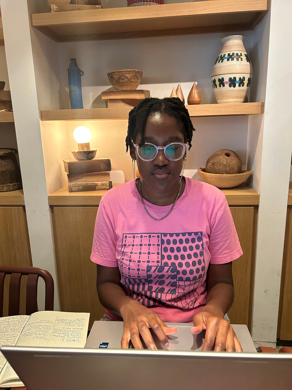 A photograph of Sojourner in a pink shirt working at a cafe table on her laptop.