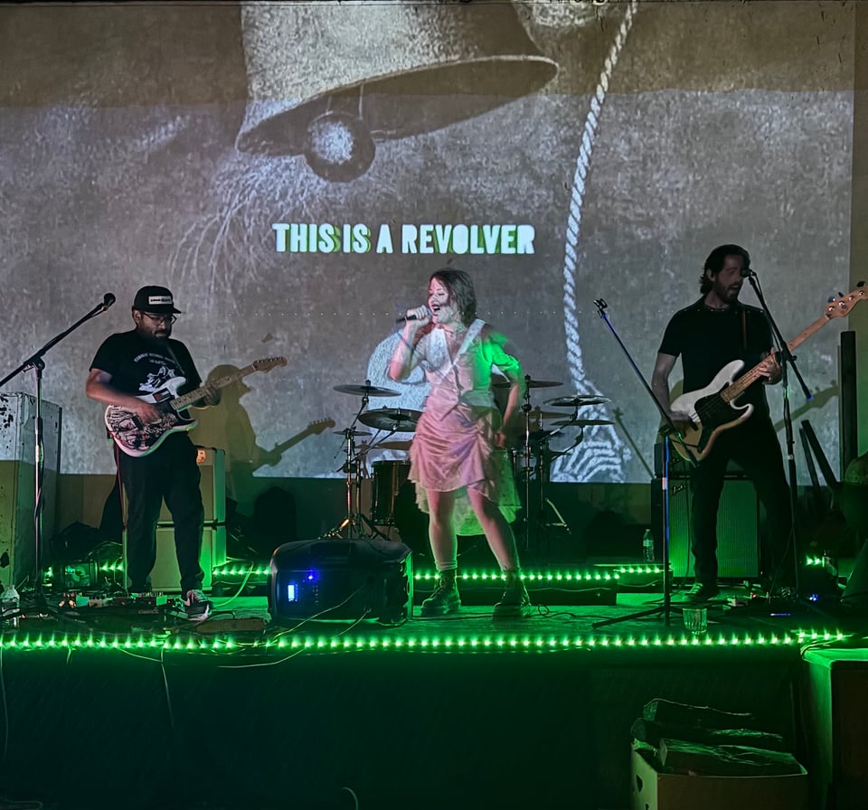 the band playing on a rickety stage lined with green strip lights. behind them is a projection of their new EP cover, which has their name, this is a revolver, below a bell. on stage rudy plays guitar, nikki sings, and matt plays bass. taylor's drums are visible behind nikki, and i promise he's there too.