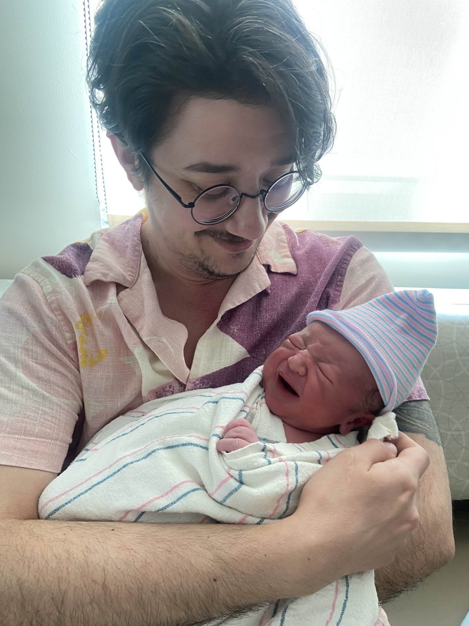 Nino, a white nonbinary person with dark hair and glasses, holding a very tiny swaddled baby. Nino gazes fondly down at the newborn, while he has his face scrunched in a cry.