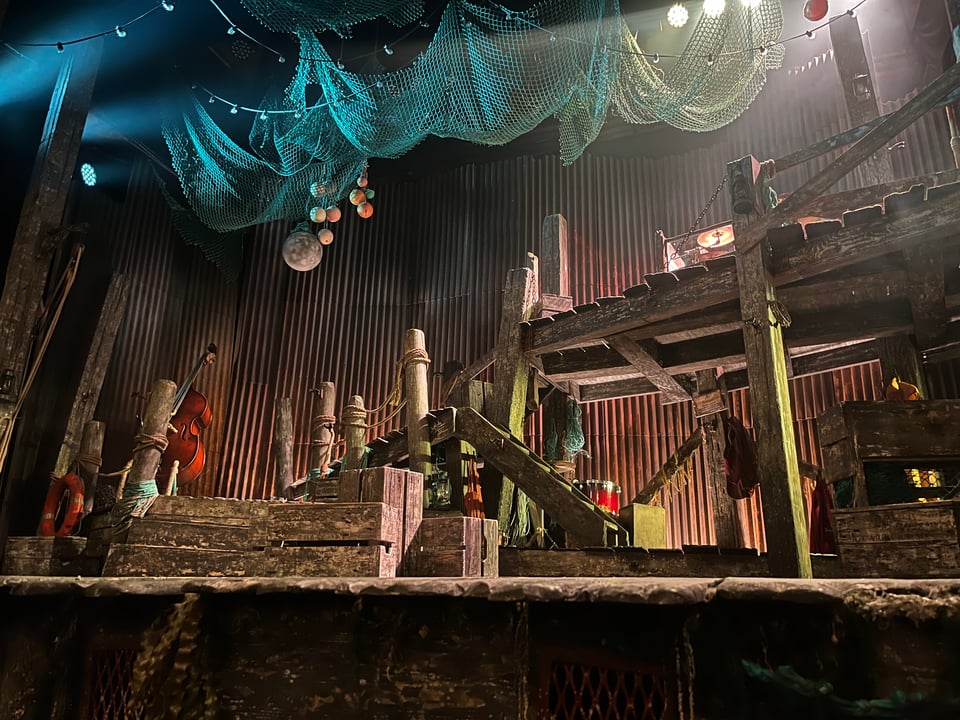 An empty theatre stage, with a set made of wood and fishing nets to conjure up the aesthetic of a Cornish fishing village.