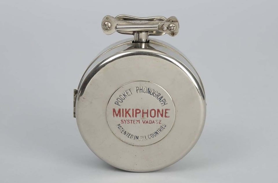 A folded up Mikiphone, which looks like a polished silver flat canister, or perhaps a pocketwatch, standing on one en