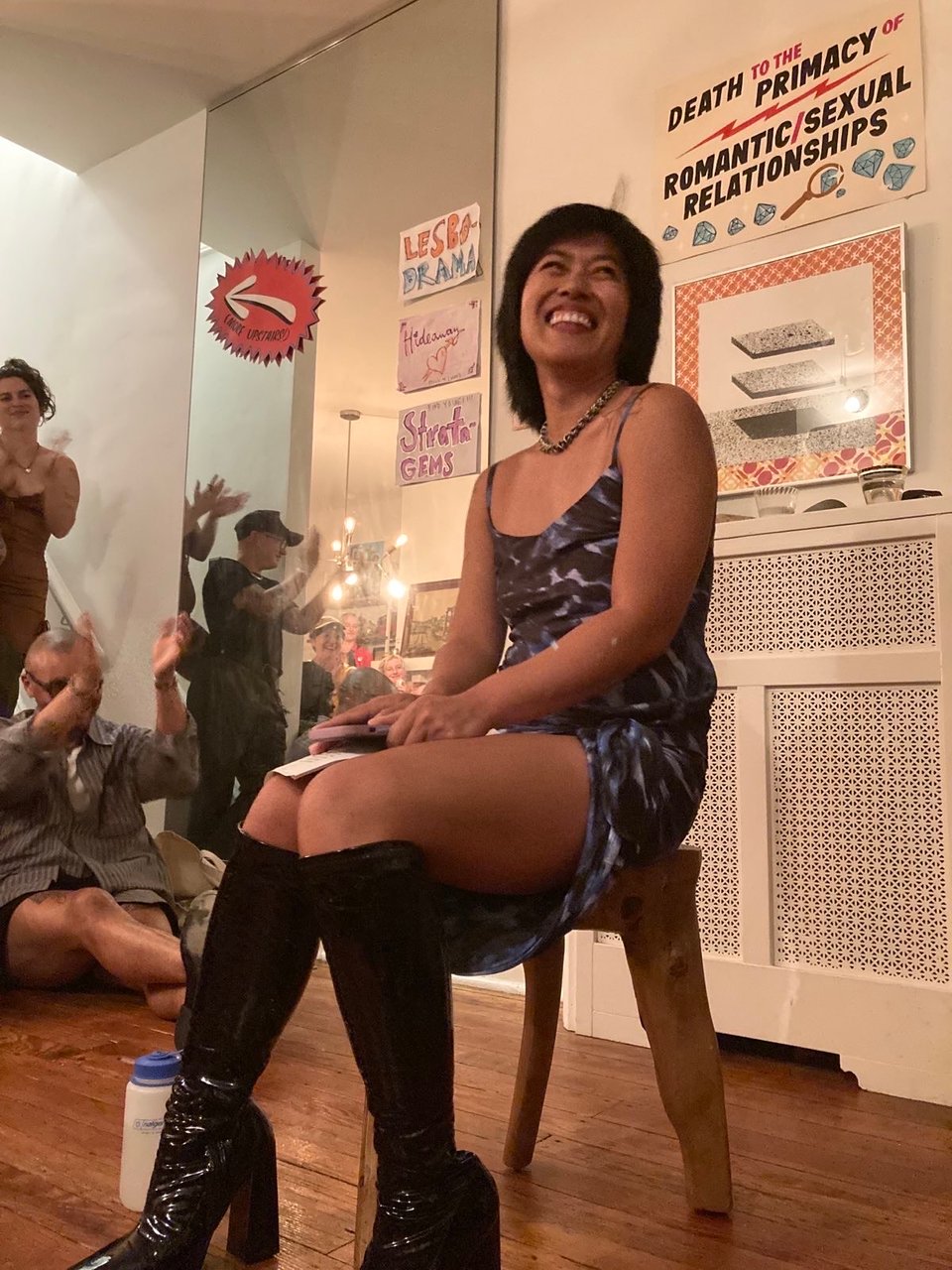 filipino femme smiles as people clap, behind her signs read "death to the primacy of romantic/sexual relationships" "lesbodrama" "stratagems"