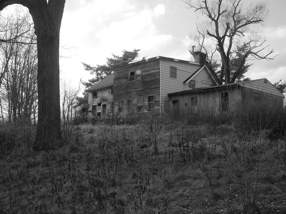 A black-and-white photo shows an abandoned, weather-beaten house on a slight hill, surrounded by leafless trees and overgrown brush. The building’s siding is warped and peeling, with boarded or broken windows hinting at long neglect. The stark contrast and barren landscape give the scene a haunting, desolate atmosphere.