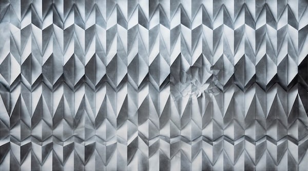 Patrón regular de triángulos y rombos con profundidades diferentes que generan un juego de sombras. Hacia media altura y ligeramente a la derecha del lienzo hay un parte de él arrugado intencionalmente.