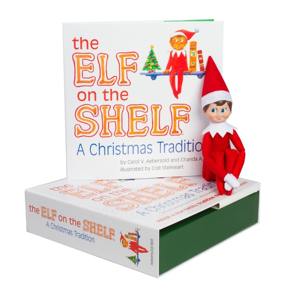 A skinny elf in red suit sits on a box in front of a book titled "the elf on the shelf: a christmas tradition"