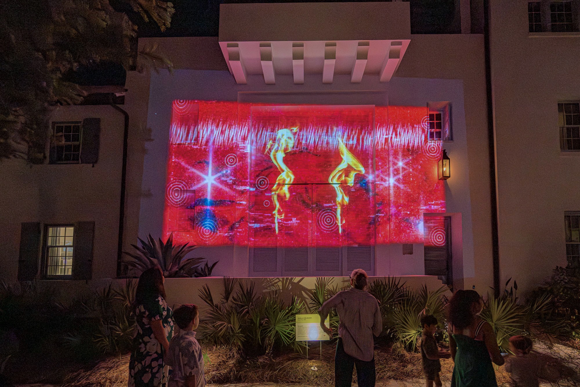 Projection on white building of two flames on red background. 3 children and 2 adults look on.