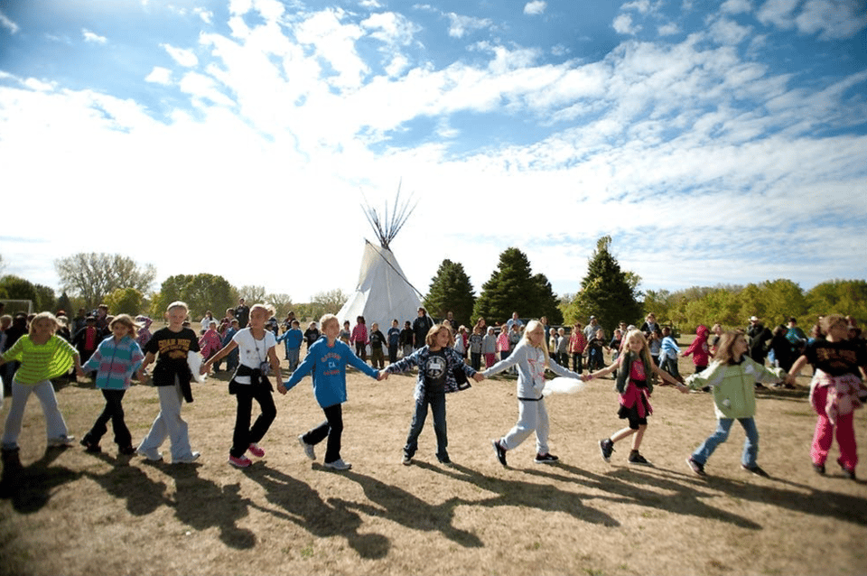 Photo of children holding hands and circling with a teepee in the background.