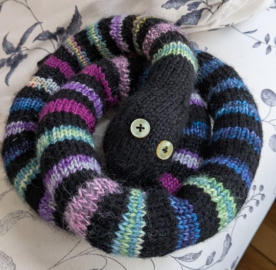 LeVar the Book Snake is curled up on the edge of a faded white floral print armchair. He has gold button eyes and many colors of stripes separated by black stripes.