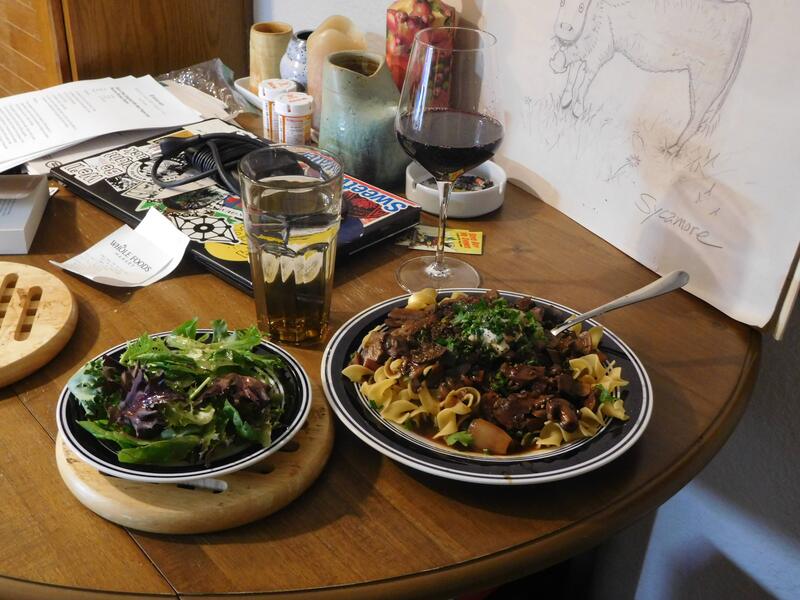 A table setting consisting of a hearty mushroom dish served over egg noodles, a plate of salad, a glass of water, and a glass of wine. The table is also cluttered with household items.