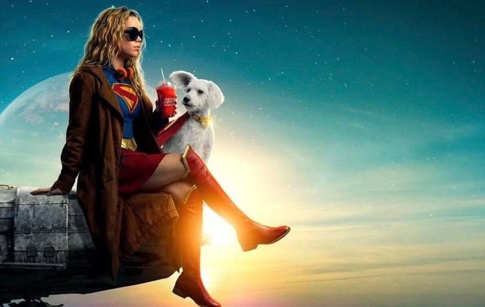 Supergirl sits on the edge of a spaceship wearing old-fashioned headphones and a brown trenchcoat over her usual superhero costume. She has a slushie in one hand and a white dog in her lap. An alien sun is going down