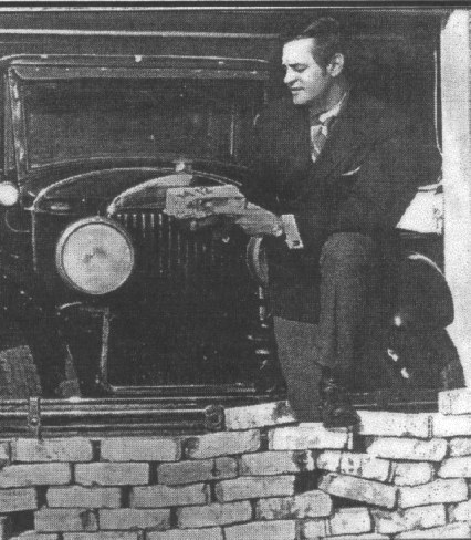 Newspapers photograph in black and white. Shows a man, George Patey, holding a brick with his foot on a partially assembled wall. Behind him is an old fashioned 1920s car.