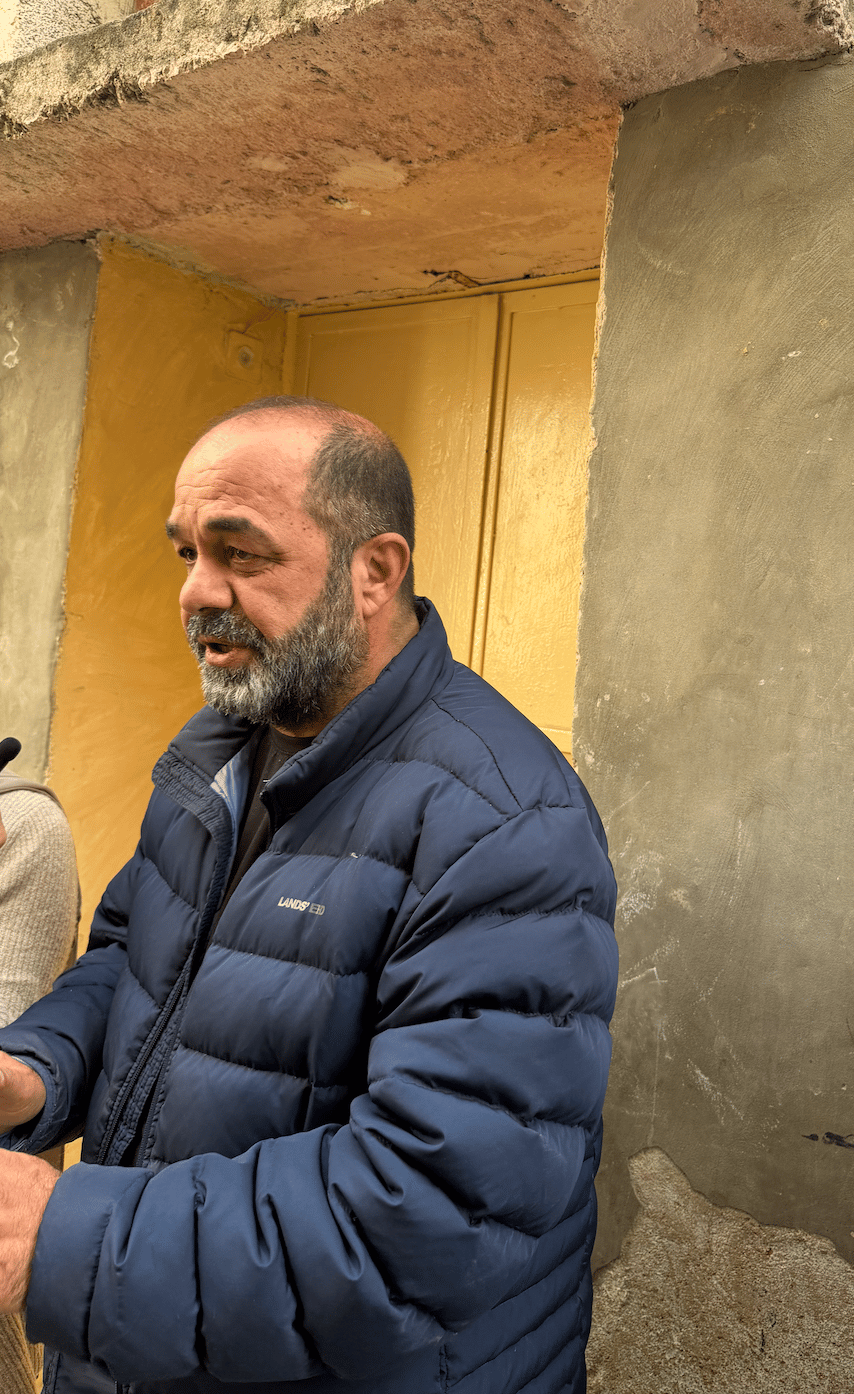 Photograph of the same man in the first photo, wearing a blue puffer jacket standing outside a yellow door next to a gray wall.