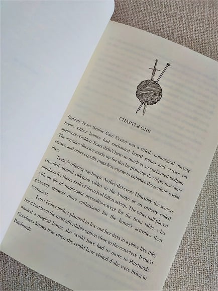 photo of a paperback of The Remarkable Retirement of Edna Fisher open to the first page, showing a black and white graphic of a yarnball stabbed through with sword and knitting needle, the heading "chapter one," and the first page of text. it's a whole book!