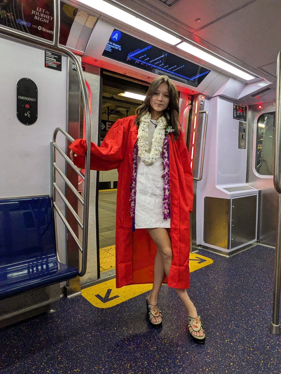 Ryo Leyh wearing a red graduation gown and a short white dress with a lei of white flowers around her neck, standing on the A train subway in NYC